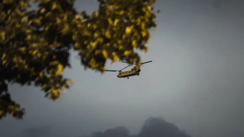 Een helikopter vliegt laag tussen bomen met gele bladeren, met op de achtergrond een berg en een grijze hemel.
