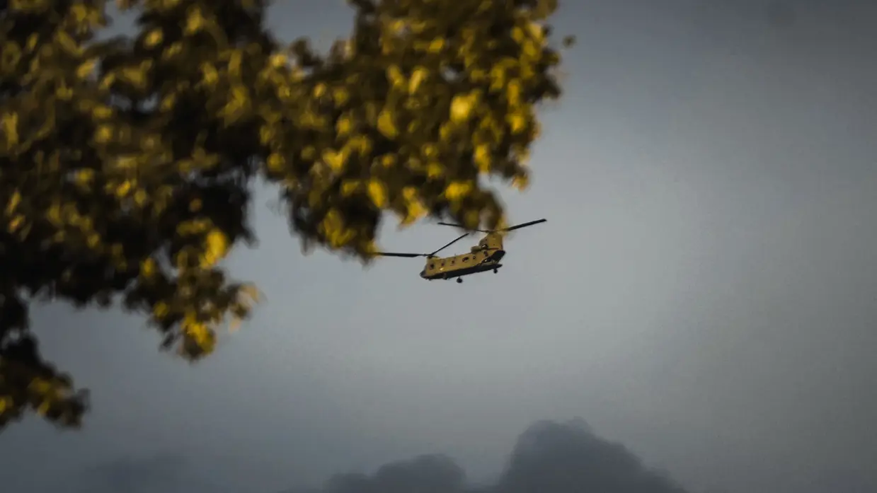 Een helikopter vliegt laag tussen bomen met gele bladeren, met op de achtergrond een berg en een grijze hemel.