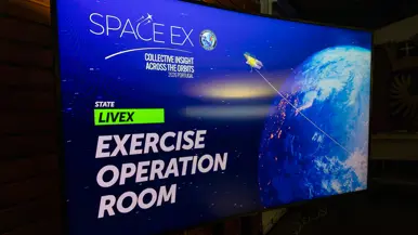 Digitaal scherm met daarop de wereldbol gezien vanuit de ruimte met daarboven een satelliet. Over de afbeelding staat de tekst: ‘SpaceEx; Collective insight across the orbits; Portugal 2026. State LiveEx; Exercise Operation Room’.