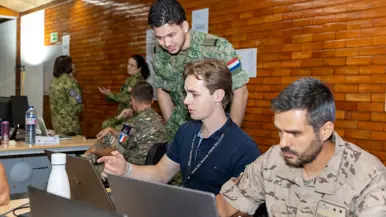 Twee militairen en een burger aan het werk op een paar laptops, een van de militairen is Nederlands. Op de achtergrond nog vier militairen aan het werk.