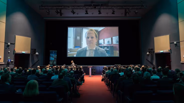 Staatssecretaris op groot scherm. Zaal mensen kijkt.