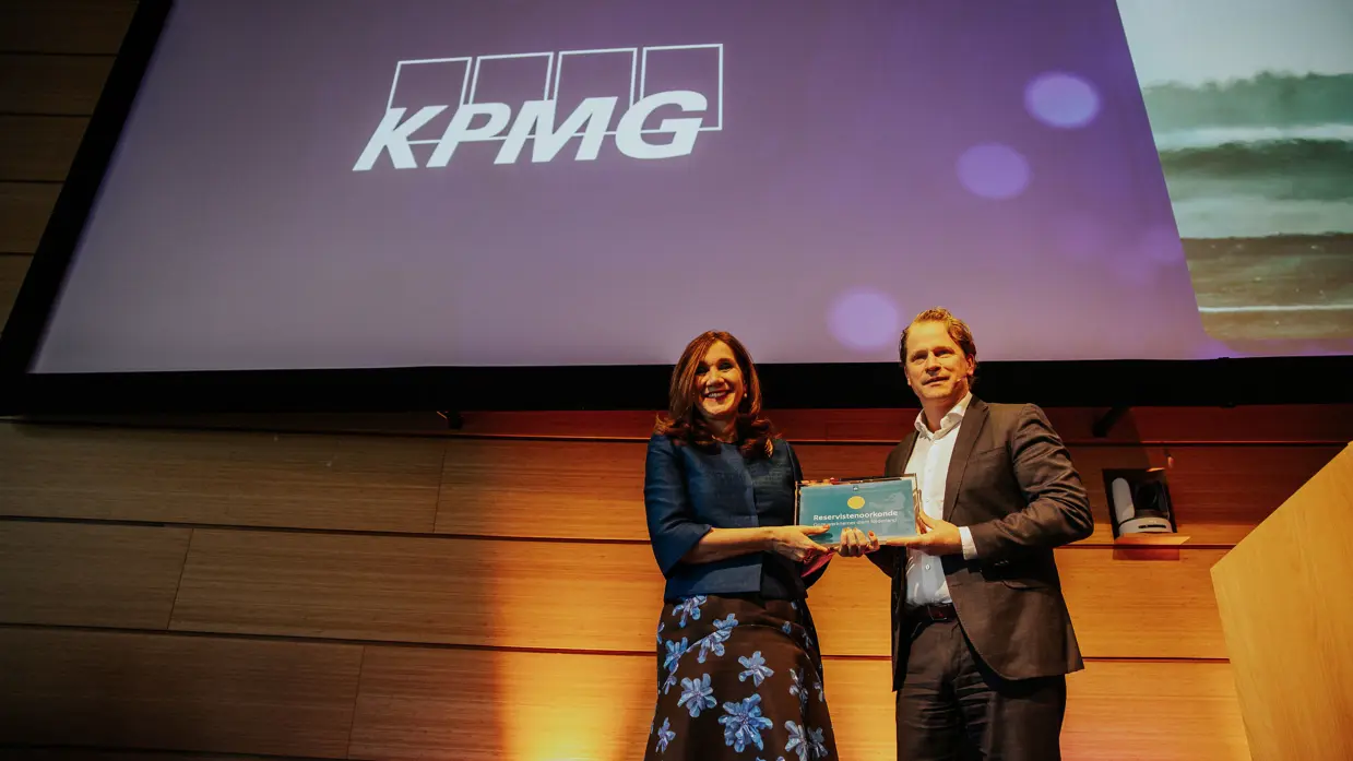 Hottenhuis en Tuinman houden samen een ingelijste oorkonde vast op een podium met boven hen een scherm met daarop de tekst ‘KPMG’.