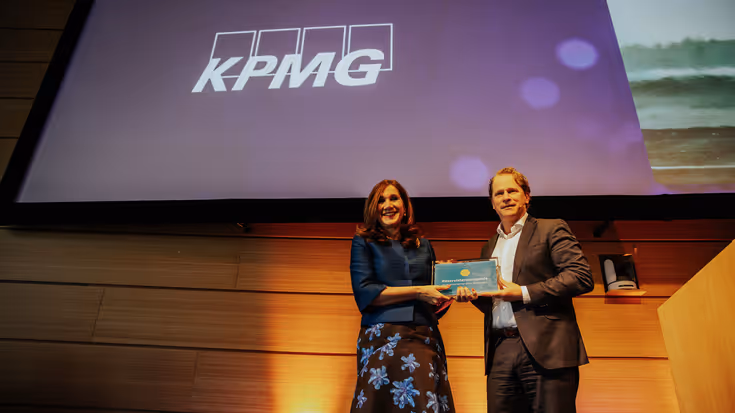 Hottenhuis en Tuinman houden samen een ingelijste oorkonde vast op een podium met boven hen een scherm met daarop de tekst ‘KPMG’.