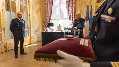  Steur staat ‘op de plaats rust’ in een chique kamer met hoog plafond. Een militair spreekt hem toe vanachter een spreekgestoelte. Een ander militair houd een bordeaux rood kussen vast met daarop een medaille.
