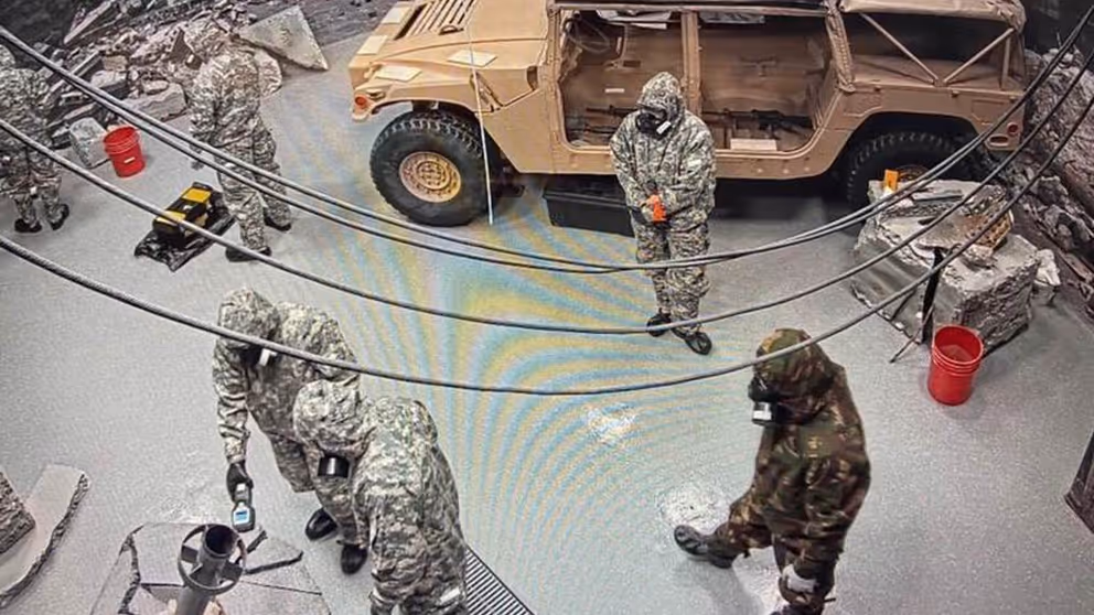 5 militairen met gasmaskers in een ruimte met een voertuig.