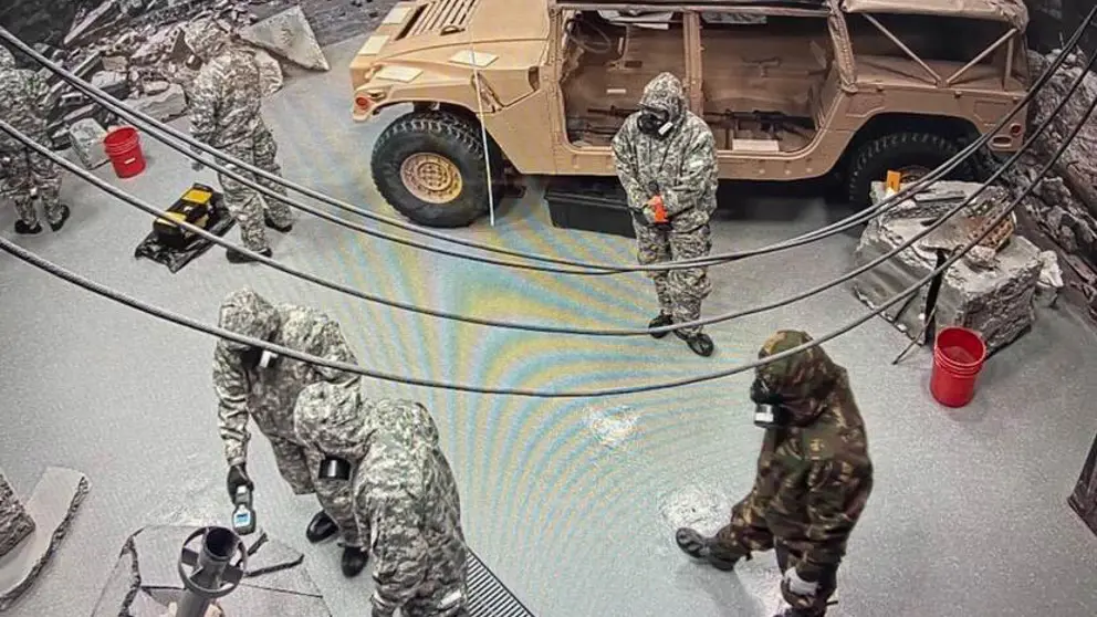 5 militairen met gasmaskers in een ruimte met een voertuig.