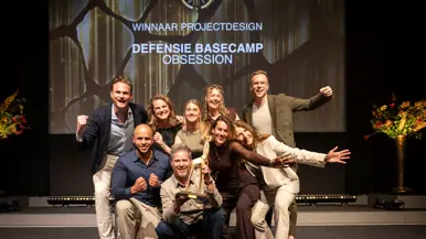 Negen mensen juichend op de foto met een award in de hand. Op de achtergrond de geprojecteerde tekst ‘winnaar projectdesign, Defensie Basecamp, Obsession’.