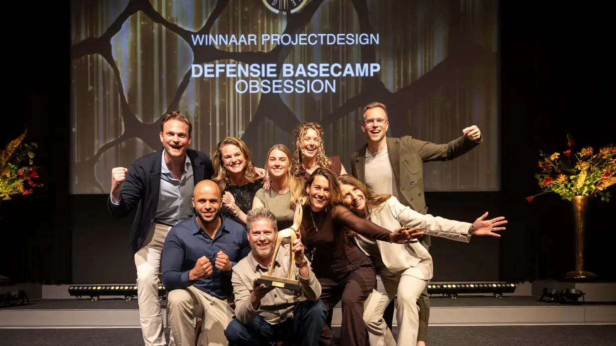 Negen mensen juichend op de foto met een award in de hand. Op de achtergrond de geprojecteerde tekst ‘winnaar projectdesign, Defensie Basecamp, Obsession’.