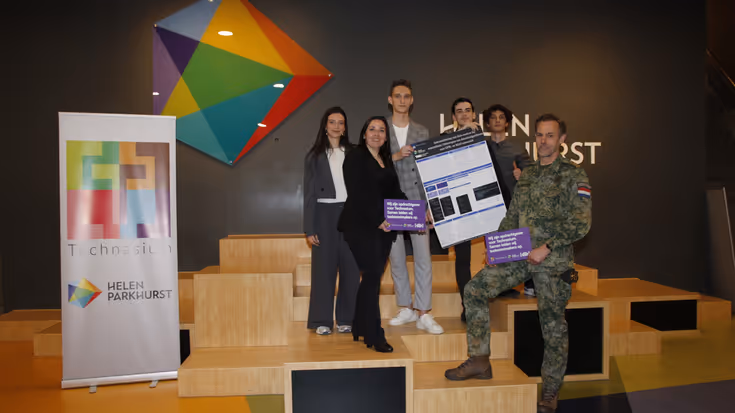 Vier scholieren poseren met een bordje in hun hand. Naast hen staan een volwassen vrouw en een mannelijke militair.
