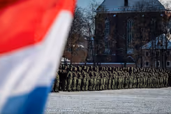 Manschappen in uniform op een rij op de achtergrond en op de voorgrond links een stukje van de Nederlandse vlag.