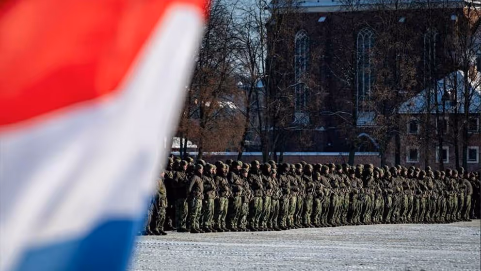 Manschappen in uniform op een rij op de achtergrond en op de voorgrond links een stukje van de Nederlandse vlag.