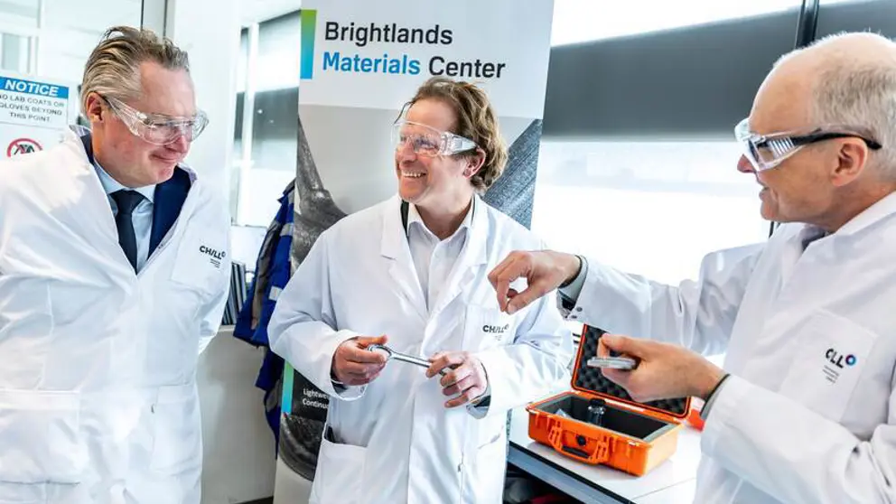 Staatssecretaris Tuinman in witte jas met veiligheidsbril praat bij de Brightlands Chemelot Campus met 2 mannen in witte jassen.