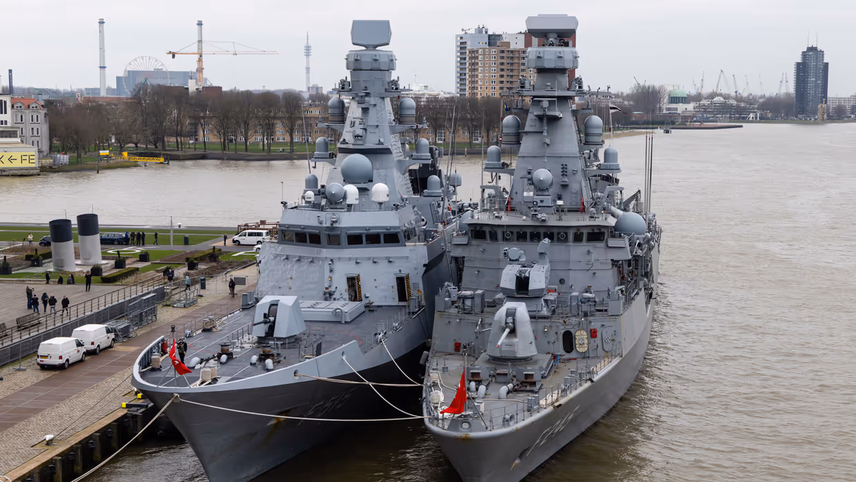 Twee oorlogsschepen liggen in een haven en op een groot schip staan helikopters opgesteld.