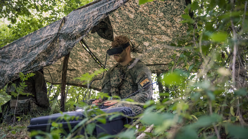 Een militair onder een gespannen zeiltje in het veld bedient de afstandsbediening van een drone en draagt een VR-bril.  