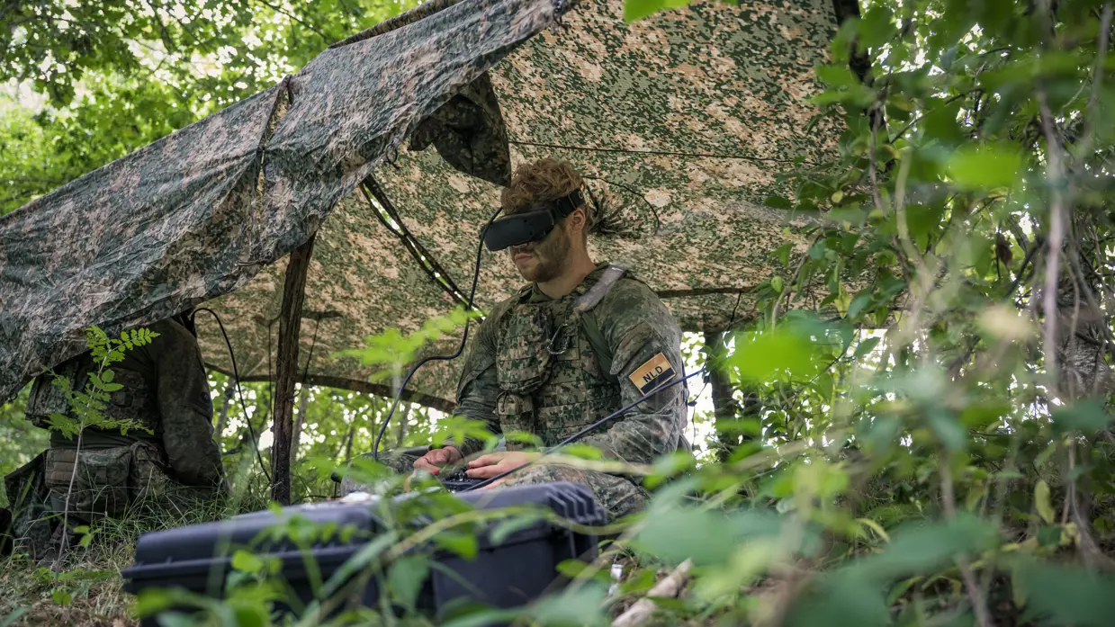 Een militair onder een gespannen zeiltje in het veld bedient de afstandsbediening van een drone en draagt een VR-bril.  