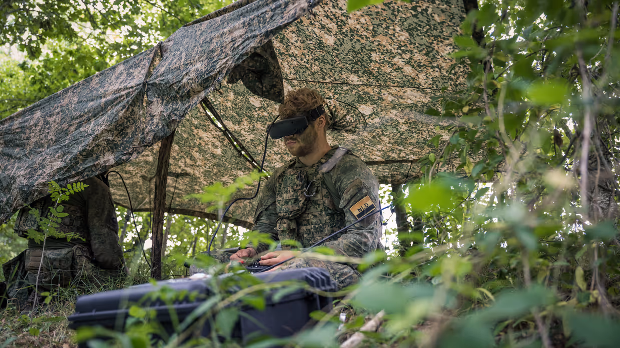 Een militair onder een gespannen zeiltje in het veld bedient de afstandsbediening van een drone en draagt een VR-bril.  