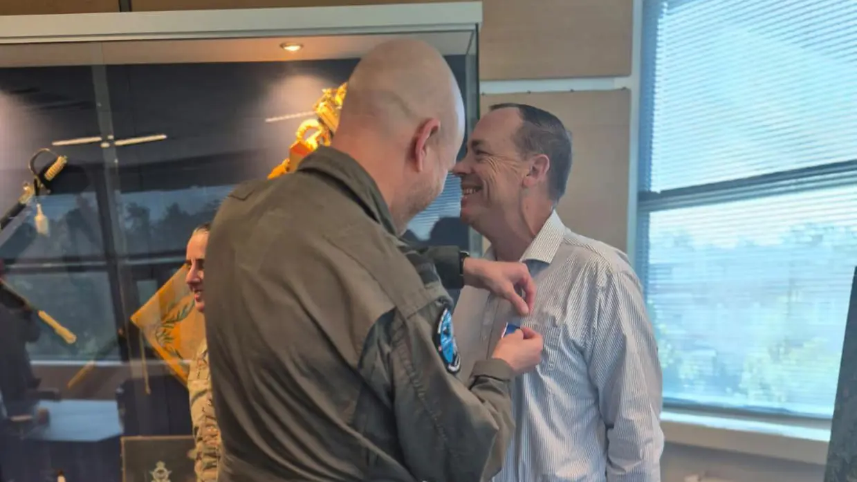 Adang spelt een medaille op de borst van Rautter. Ernaast staat Lee in de houding. Ze staan op een blauw tapijt met daarop een herhaalde print van het luchtmachtembleem.