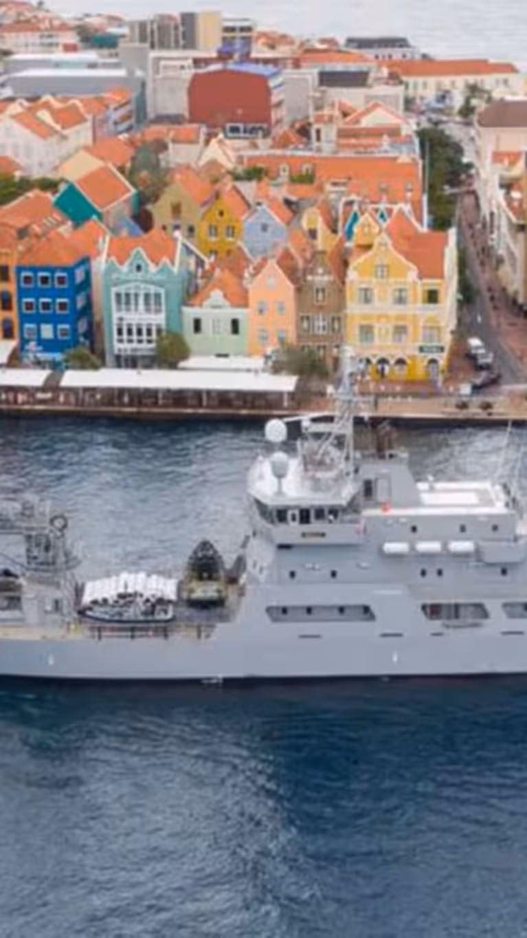 Een grijs marineschip vaart op kalm water langs kleurrijke, lage gebouwen aan de kust van een levendige stad.