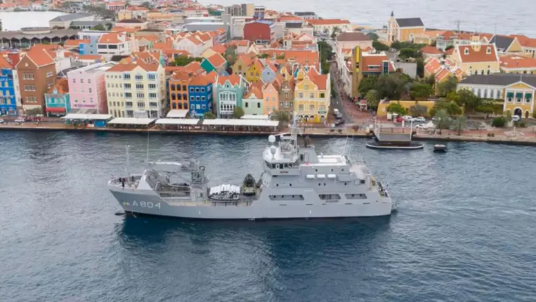 Een grijs marineschip vaart op kalm water langs kleurrijke, lage gebouwen aan de kust van een levendige stad.