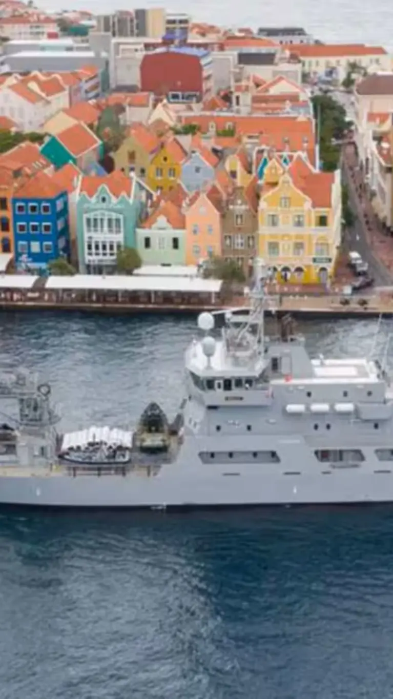 Een grijs marineschip vaart op kalm water langs kleurrijke, lage gebouwen aan de kust van een levendige stad.