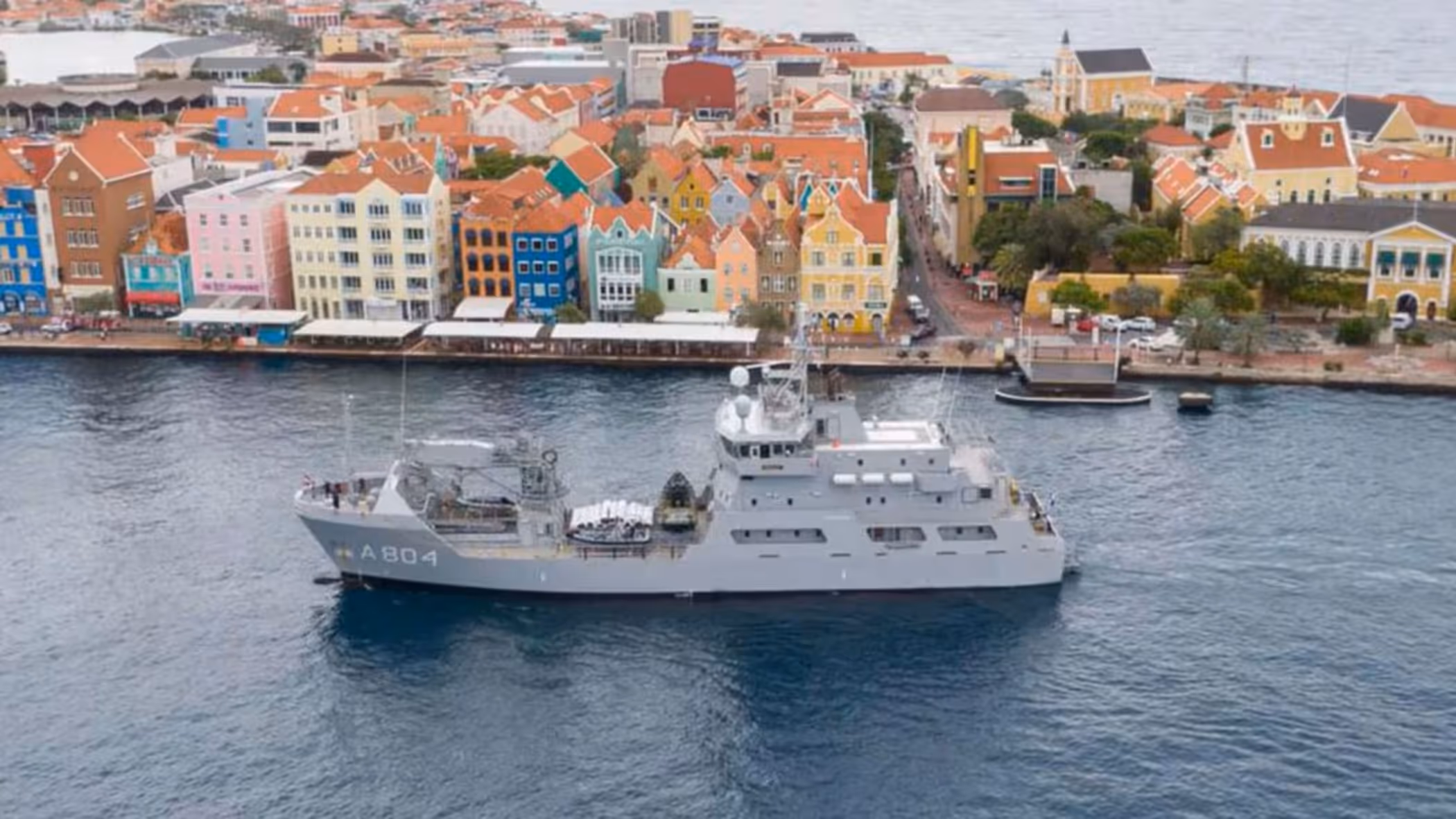 Een grijs marineschip vaart op kalm water langs kleurrijke, lage gebouwen aan de kust van een levendige stad.