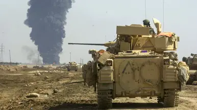 Links: Een rij militaire pantservoertuigen rijdt op weg naar Bagdad.Boven de weg staan verkeersborden met Mosul, Abu Ghraib en White Gold Village. Er zijn vlaggen te zien in een stoffig landschap. Rechts: Een pantserwagen staat op een stoffig, open veld. Op de achtergrond stijgt een dikke, zwarte rookkolom omhoog.  Soldaten en uitrusting zijn zichtbaar rondom het voertuig.