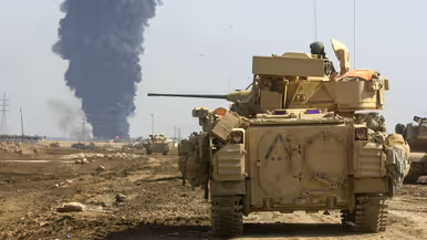 Links: Een rij militaire pantservoertuigen rijdt op weg naar Bagdad.Boven de weg staan verkeersborden met Mosul, Abu Ghraib en White Gold Village. Er zijn vlaggen te zien in een stoffig landschap. Rechts: Een pantserwagen staat op een stoffig, open veld. Op de achtergrond stijgt een dikke, zwarte rookkolom omhoog.  Soldaten en uitrusting zijn zichtbaar rondom het voertuig.