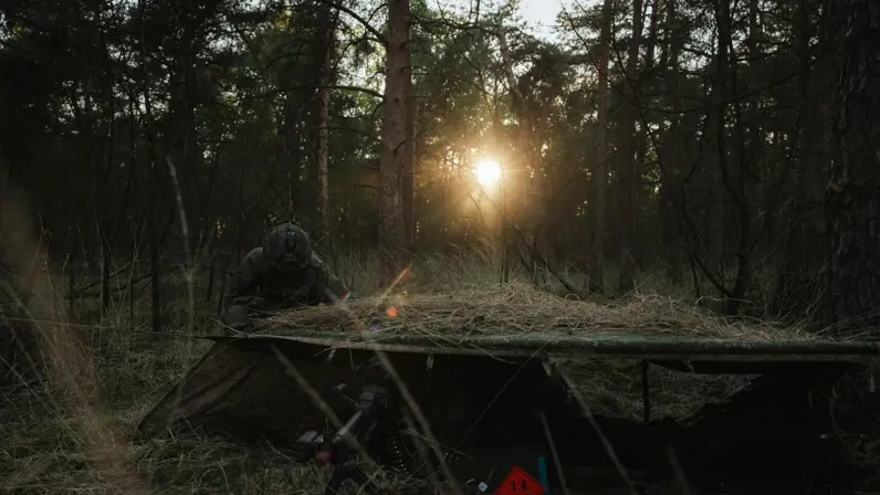 Een militair ligt in stelling naast een gecamoufleerde positie in een bosgebied terwijl de zon opkomt tijdens oefening Versatile Guard.