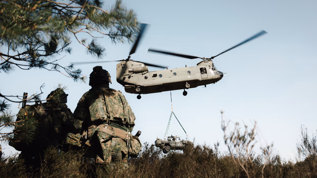 Militairen kijken toe terwijl een Chinook een voertuig onder de romp vervoert tijdens oefening Versatile Guard. 