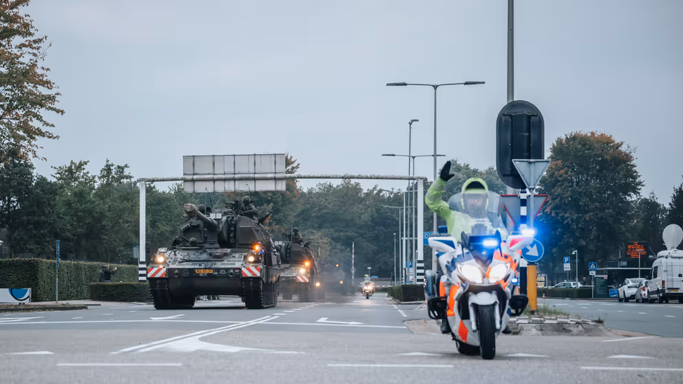 Een militaire colonne rijdt over de weg, daarvoor een motor van de Koninklijke Marechaussee.