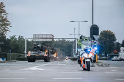 Een militaire colonne rijdt over de weg, daarvoor een motor van de Koninklijke Marechaussee.