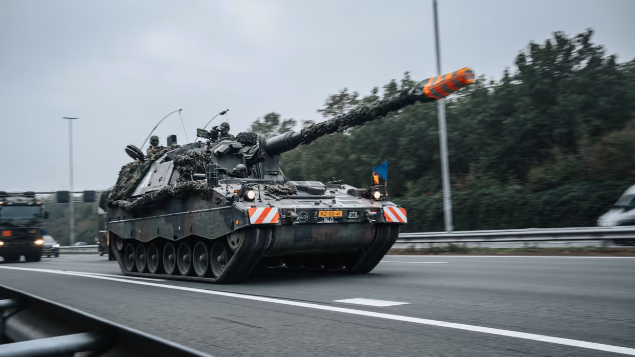 Een Pantserhouwitser rijdt over de snelweg, daarachter een vrachtwagen.