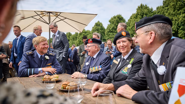 Een lachende koning Willem-Alexander aan een picknicktafel in gesprek met een veteraan.