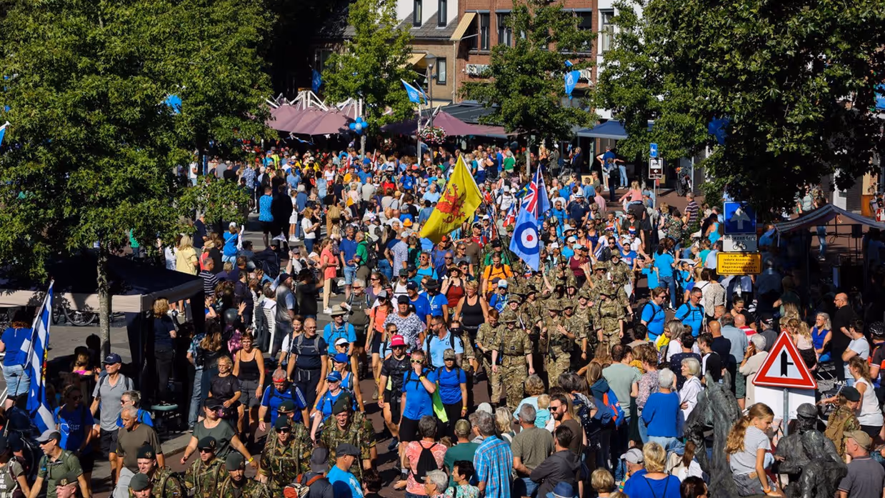 Militairen en wandelaars van de Vierdaagse lopen door straat met rijen publiek die hen aanmoedigen.