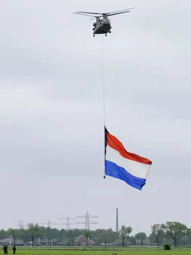 Een Chinook hangt stil boven een weiland met eronder een enorme Nederlandse vlag. Links onder staan drie mensen. Midden onderin staan een boerderij, een paar huizen, hoogspanningsmasten en een hoge schoorsteen.