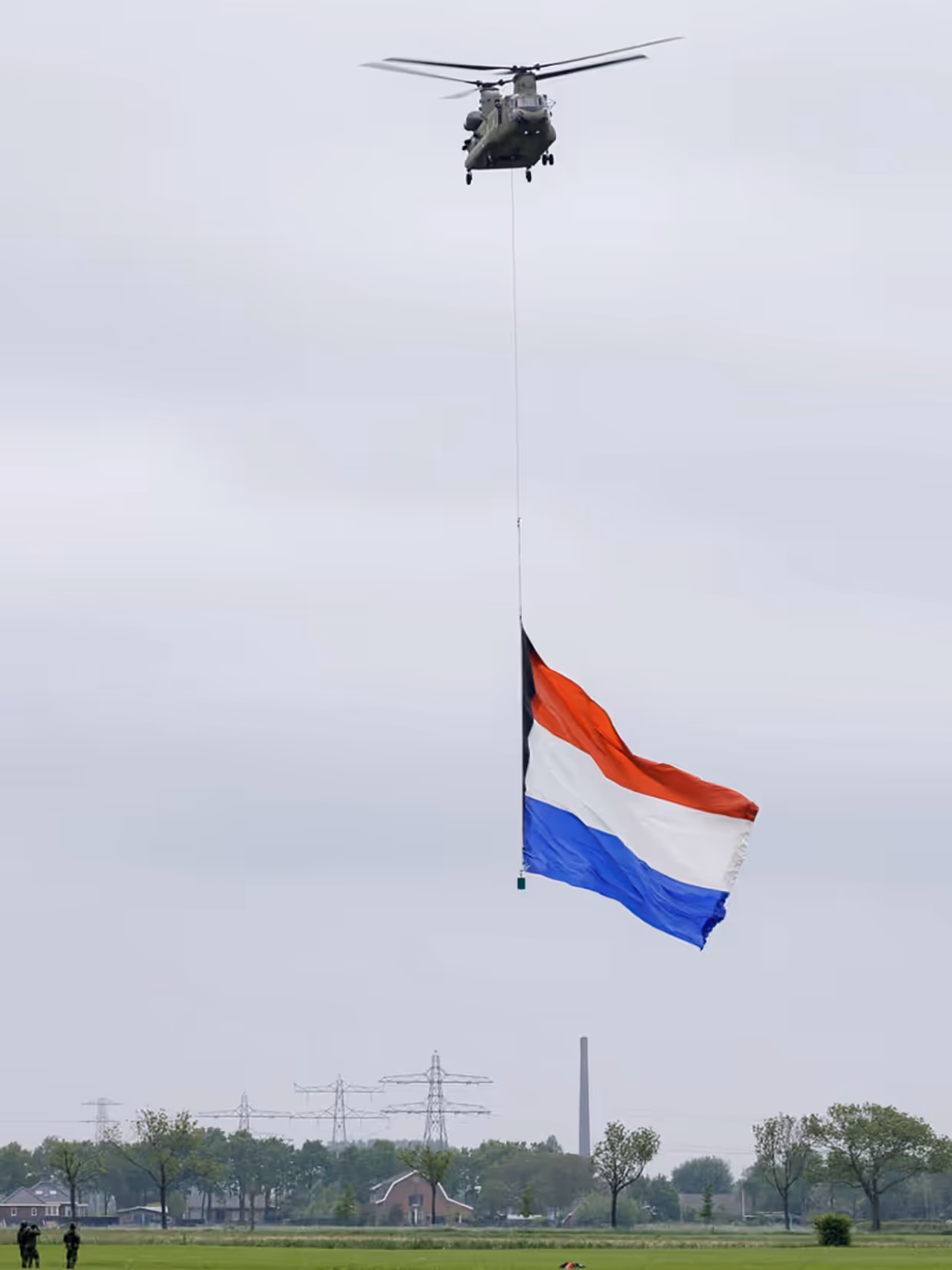 Een Chinook hangt stil boven een weiland met eronder een enorme Nederlandse vlag. Links onder staan drie mensen. Midden onderin staan een boerderij, een paar huizen, hoogspanningsmasten en een hoge schoorsteen.