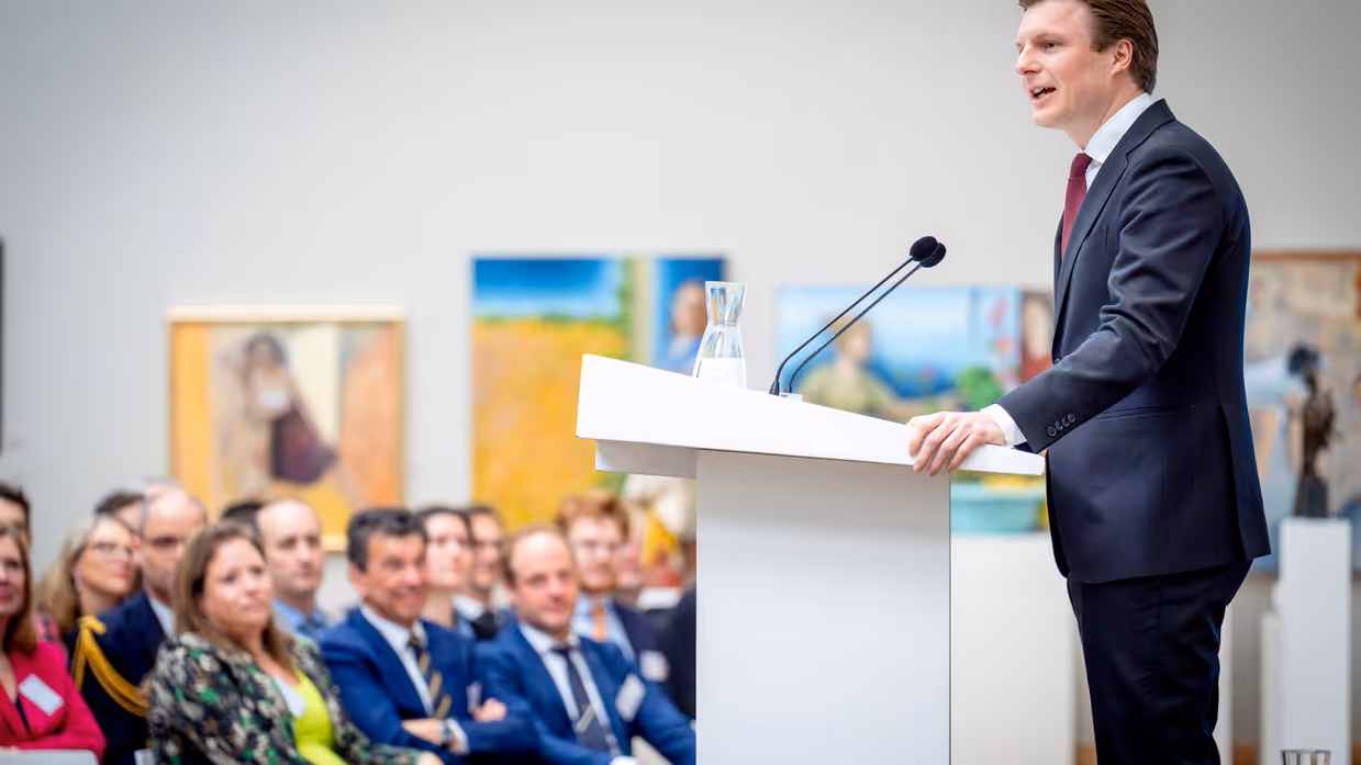 Man in pak achter katheder spreekt zaal toe.