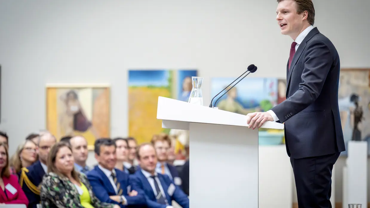 Man in pak achter katheder spreekt zaal toe.