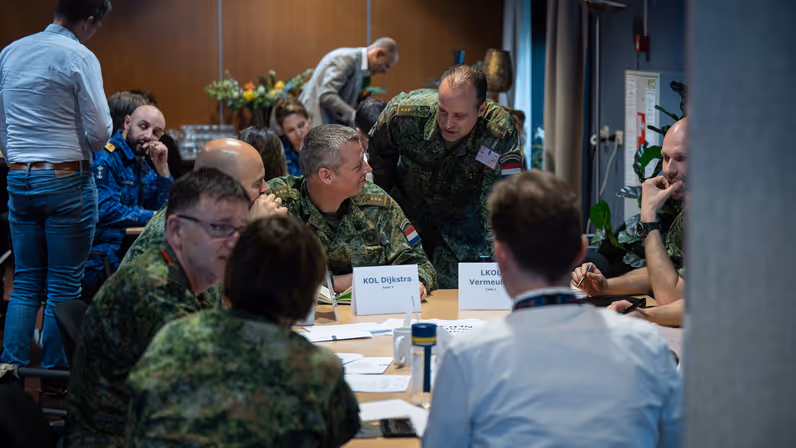 Militairen van land- en luchtmacht en marine, samen met burgers in overleg aan twee ronde tafels.