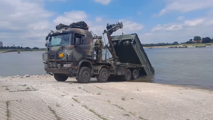 Een militaire vrachtwagen leegt de laadbak in een rivier.