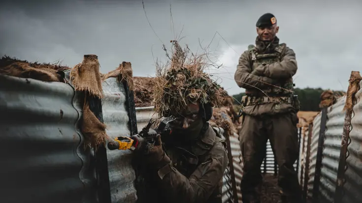 Een militair staat met zijn armen over elkaar. Hij kijkt mee over de schouders van een collega die oefent met een wapen in een loopgraaf.