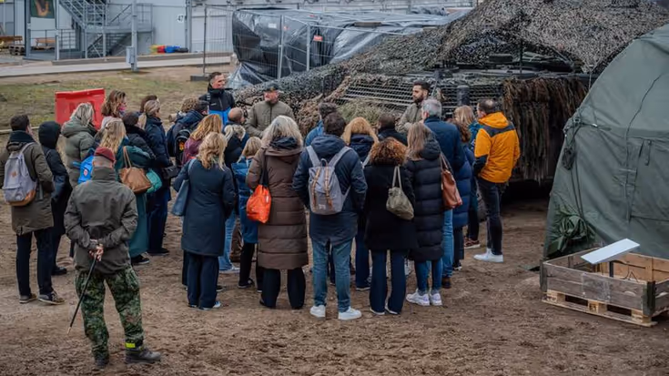 Mensen in burger luisteren naar militair voor de groep. Op de achtergrond militair materieel.