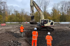 Hijskraan bij afgraving grond. 3 mensen met witte helm in oranje pakken staan erbij.