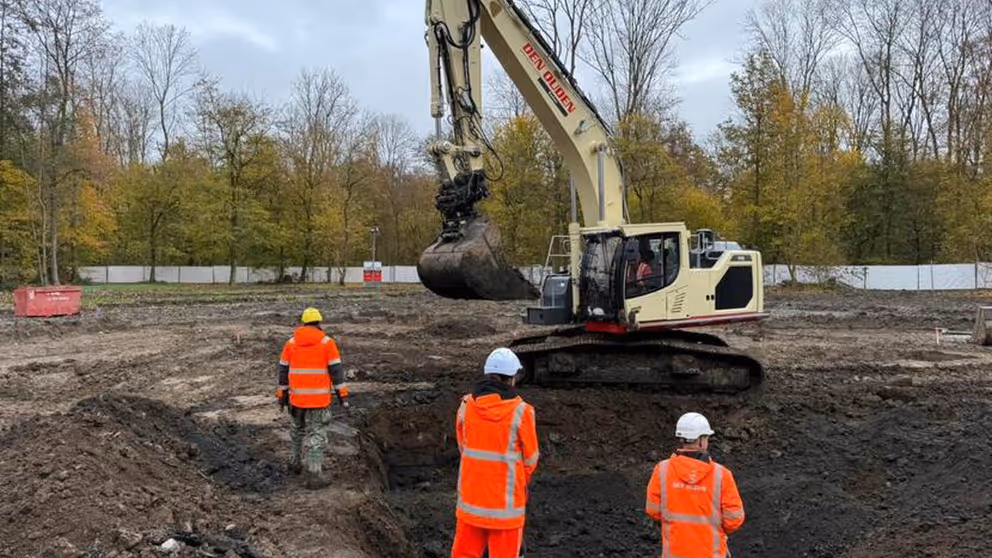 Hijskraan bij afgraving grond. 3 mensen met witte helm in oranje pakken staan erbij.