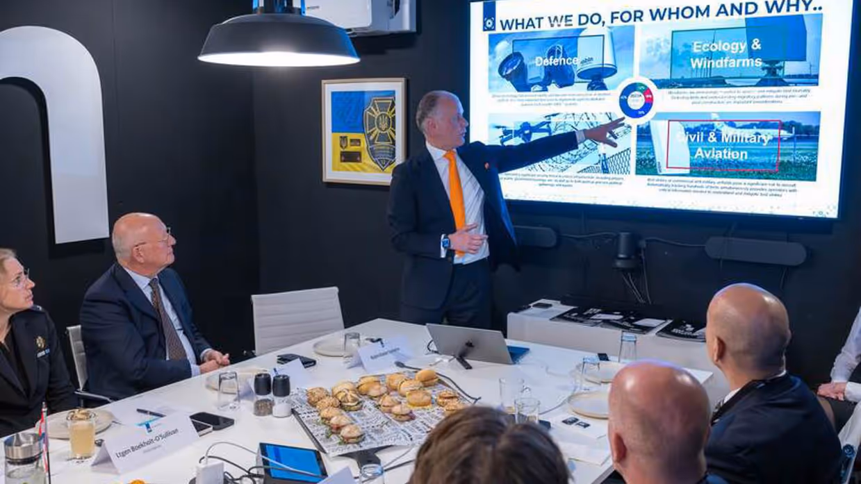 Tafel waar mensen aanzitten. Een staat en wijst naar groot scherm.