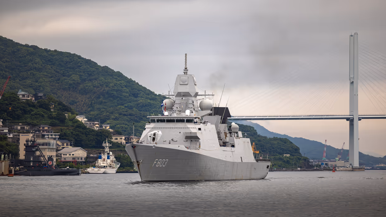 Een varend, grijs marineschip. Op de achtergrond een brug en groene heuvels.